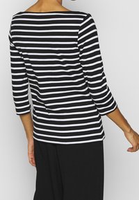 Persona che indossa una maglietta a maniche lunghe a righe orizzontali bianche e nere e pantaloni neri, vista di spalle contro uno sfondo semplice.