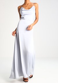 Kvinna klädd i en ärmlös, ljusblå satin-klänning med tunna axelband och svarta klackskor med öppen tå, stående mot en enkel vit bakgrund.