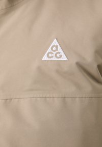 Beige fabric with a smooth texture features a white embroidered triangle logo, centered, with a geometric design.