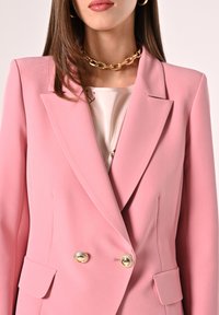 Rosa übergroße Blazer mit doppelreihigen goldenen Knöpfen, scharfen Revers und glatter Textur. getragen über einer hellen Bluse.