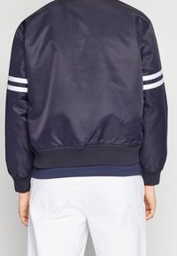 Námořnická modrá bomber bunda vyrobená z hladkého nylonu s bílými pruhy na rukávech, žebrovanými manžetami a lemem, vystihující volný střih.