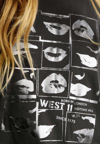 Camiseta negra con un patrón de cuadrícula de labios blancos y el texto "WEST 11 BORN IN LONDON NOTTING HILL SINCE 1973" en negrita.