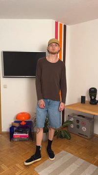 Rjava majica z dolgimi rokavi, modri jeans kratke hlače, črne čevlje z lahkimi podplati in črne nogavice. Stoji v prostoru z barvitim dekorjem.