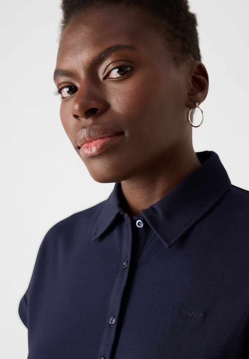 Granatowa koszulka polo z teksturowanej tkaniny, kołnierzykiem i zapięciem na guziki. Haftowane logo "GANT" po lewej stronie. Srebrny kolczyk w kształcie kółka.