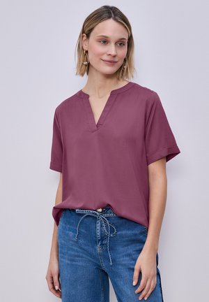 Vrouw draagt een paarse blouse met korte mouwen en V-hals en een blauwe spijkerbroek met een koordriem, staand tegen een effen lichte achtergrond.