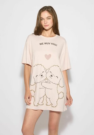 Młoda kobieta nosząca blado różową oversize koszulę z ilustracjami białego szczeniaka przytulającego się oraz tekstem "WE WUV YOU!" nad sercem.