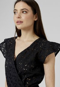 Robe noire avec un motif en dentelle florale, manches courtes flottantes, décolleté en V et tissu texturé. Les détails incluent des découpes ouvertes sur toute la robe.