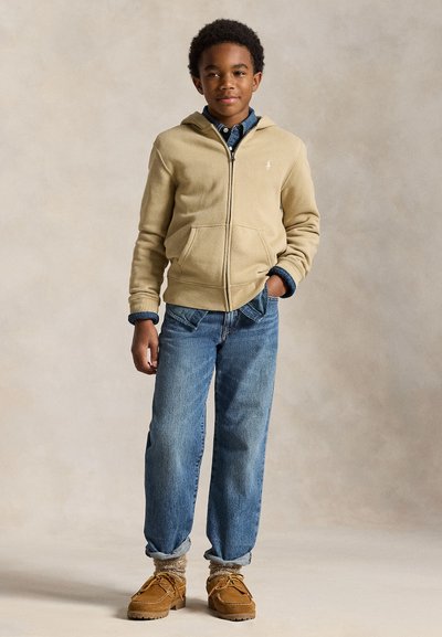 Jongen staat met één hand in zijn zak, draagt een beige zip-up hoodie, een blauw overhemd, opgerolde spijkerbroek, dikke sokken en bruine schoenen