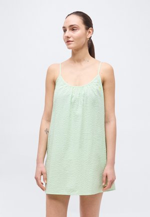 Mladá žena so zviazanými vlasmi, oblečená v svetlozelených šatách bez rukávov s tenkými ramienkami, stojaca pred jednoliatym pozadím.