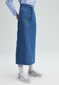 Jupe maxi en denim à taille haute, en tissu bleu, avec des rivets en argent dispersés sur toute la surface et une fermeture à boutons sur le devant. Associée à des baskets grises.