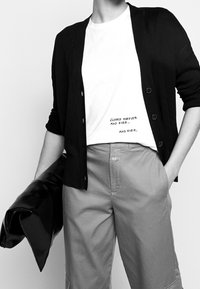 Sort strikket cardigan med knapper, hvid t-shirt med trykt tekst og beige bukser med lommer, der holder en sort lædertaske.
