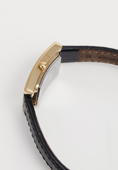 Montre dorée avec un boîtier rectangulaire, un bracelet en cuir noir et une couture décorative. Elle présente une couronne proéminente et un dos en ton argent.