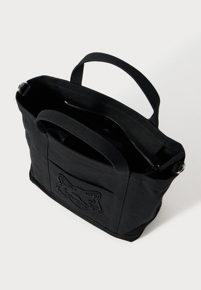 Čierna látková taška na rameno s dvoma rúčkami, predným vreckom s vyšívaným zvieracím tvárou a čiastočne otvoreným zipsom na vrchu.