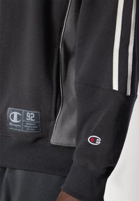 Sweatshirt preto com detalhes cinzentos, apresentando riscas brancas, um logo da Champion e punhos canelados para um design ajustado.