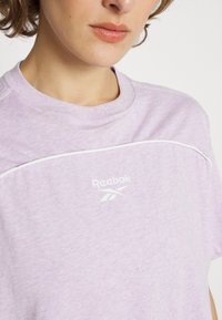 Jasnofioletowa koszulka Reebok z krótkim rękawem, okrągłym dekoltem, białym logo i białym wykończeniem wzdłuż szwów ramion. Miękka tekstura.