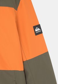 Orange och olivgrön regnjacka med en slät textur, färgblockdesign och en svart logotypklistermärke på ärmen.