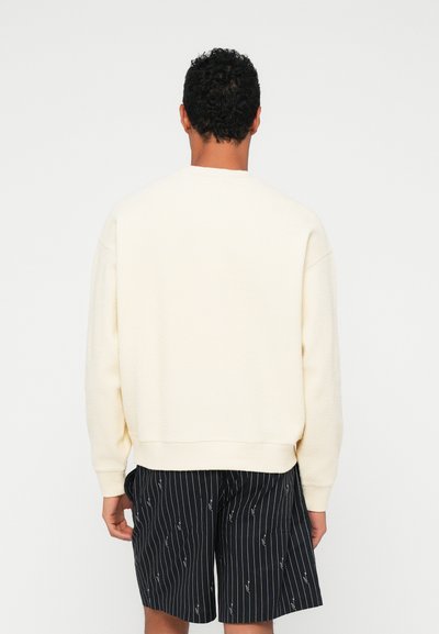Sweatshirt texturé de couleur crème avec un col rond et une coupe décontractée, associé à un short noir à rayures fines présentant un motif subtil.