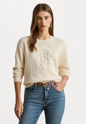 Cream knit sweater featuring an embroidered logo, round neckline, and ribbed cuffs; paired with blue jeans and a brown belt.