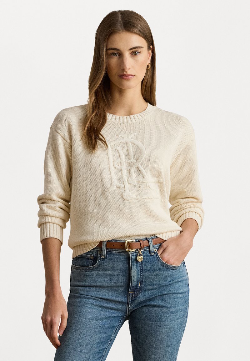 Pull en tricot crème avec logo brodé, col rond et poignets côtelés ; associé à un jean bleu et une ceinture marron.