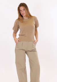 Sieviete, kas valkā spīdīgu bēša krāsas īsroku džemperi un bēša krāsas platām kājām bikses ar augstu jostasvietu, rokas ielikusi kabatās, stāvot pret balto fonu.