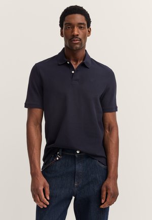 Mann in einem dunkelblauen Poloshirt und blauen Jeans, der an einem hellen, einfarbigen Hintergrund steht, mit entspannten Händen an den Seiten.