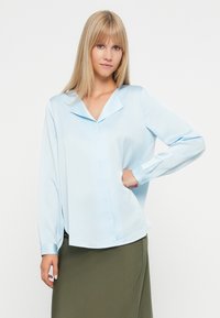 Svetlo modra bluzica s koničastim ovratnikom, dolgimi rokavi in zapenjanjem na gumb. Gladek material z udobnim krojem. Poudarjena z globokimi manšetami.