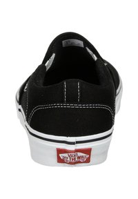 Scarpe slip-on nere in tela con suola in gomma bianca. La vista posteriore mostra delle cuciture bianche e una patch rossa con il logo "Off the Wall". Design classico.