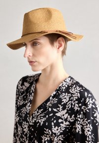 Sombrero de paja trenzada marrón con ala ancha y una banda de cuero marrón. La modelo lleva una blusa de patrón floral en blanco y negro. Fondo minimalista.