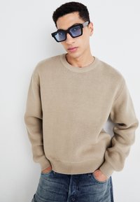 Beige striktrøje med rund halsudskæring og ribbede manchetter. Båret med mørkeblå denim jeans. Store, kantede sorte solbriller med blå linser.