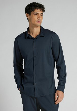 Junger Mann mit kurzen lockigen Haaren, der ein dunkelblaues langärmliges Hemd mit Knöpfen trägt, steht mit einer Hand in der Tasche vor einem schlichten Hintergrund.