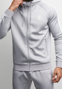 Felpa con cappuccio grigia con zip, realizzata in tessuto morbido con una texture liscia. Presenta tasche laterali, polsini a coste e un logo bianco sul petto.