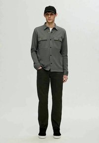 Graues Hemd mit Knöpfen und zwei Fronttaschen, schwarze Jeans und eine schwarze Mütze. Das Hemd hat eine glatte Textur und eine taillierte Passform.