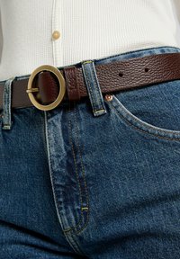 Cinturón de cuero marrón con una hebilla de latón redonda, usado sobre unos jeans de mezclilla azul. Los jeans tienen costuras amarillas y un acabado texturizado.