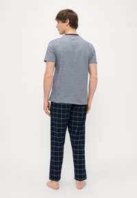 Chemise grise à manches courtes avec un col noir. Pantalon à carreaux bleu marine avec des accents jaunes. Le modèle est pieds nus, de dos, montrant l'arrière de la tenue.