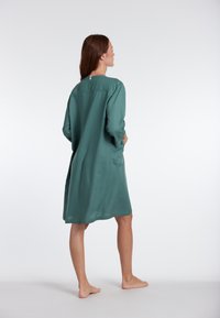 Turquoise groene jurk, tot de knie met lange mouwen, gladde textuur, sluiting met knopen op de rug en zijzakken, blootsvoets gedragen.
