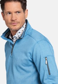 Lichtblauwe rits-sweater met een hoge kraag, textuurstof en een ritsvak op de linker mouw met gestreepte accenten.