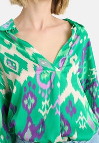 Bluzka w zielono-kremowy wzór z fioletowymi akcentami; ma dekolt w serek, kołnierzyk i zwiewną teksturę materiału.