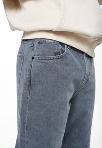 Šedé džíny z denimu se rovnýma nohama, s texturovaným povrchem, viditelným prošíváním a malou nášivkou poblíž pásku.