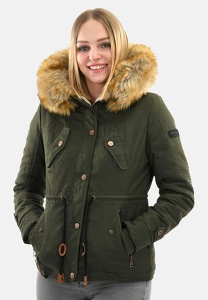 Smiling woman wearing a green winter jacket with large fur-trimmed hood and hands in pockets, standing against a light background.