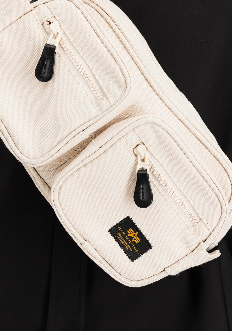 Beżowa torebka na pasek z tkaniny z dwiema zapinanymi kieszeniami, czarnymi suwakami i czarną metką z żółtym logo. Teksturowany, kompaktowy design.