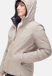 Giacca imbottita grigio chiaro con fodera interna nera, dettaglio con cerniera arancione e cappuccio. Presenta tasche laterali e un logo sul petto.