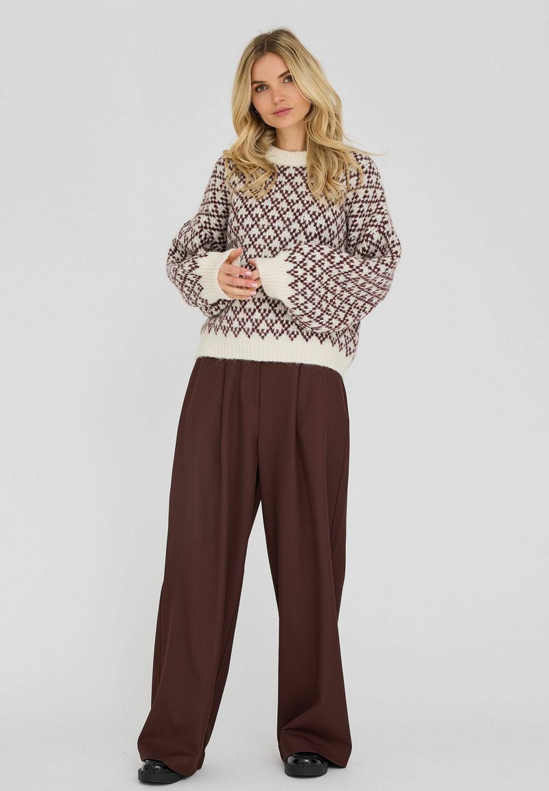 Femme aux longs cheveux blonds portant un pull en tricot à motifs et un pantalon large marron foncé, debout, les mains jointes devant elle.