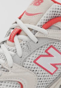 Närbild av en vit och röd New Balance löparsko med meshmaterial, vita snören och röda logodetaljer på sidan och på tungan.