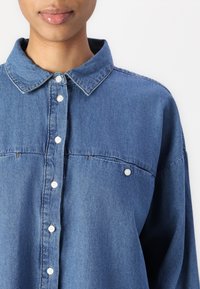 Kék denim ing, gombos elülső résszel, két mellzsebbel és klasszikus gallérral. Lazább fitetésű, valamint látható varrási részletekkel rendelkezik.