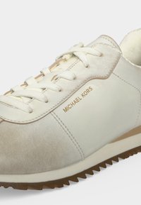 Λευκό και μπεζ αθλητικό παπούτσι Michael Kors με κορδόνια και ραβδωτή καφέ σόλα, με το όνομα της μάρκας στην πλαϊνή πλευρά.