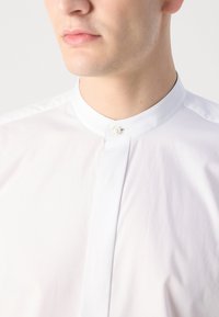 Camisa de cuello blanco con cierre de botones, fabricada en un tejido suave; cuenta con un cuello recto y un diseño limpio y minimalista.