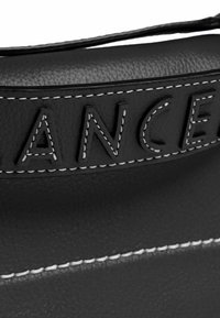 Sac en cuir noir avec surface texturée, présentant des lettres en relief et des coutures blanches contrastées le long de la couture.