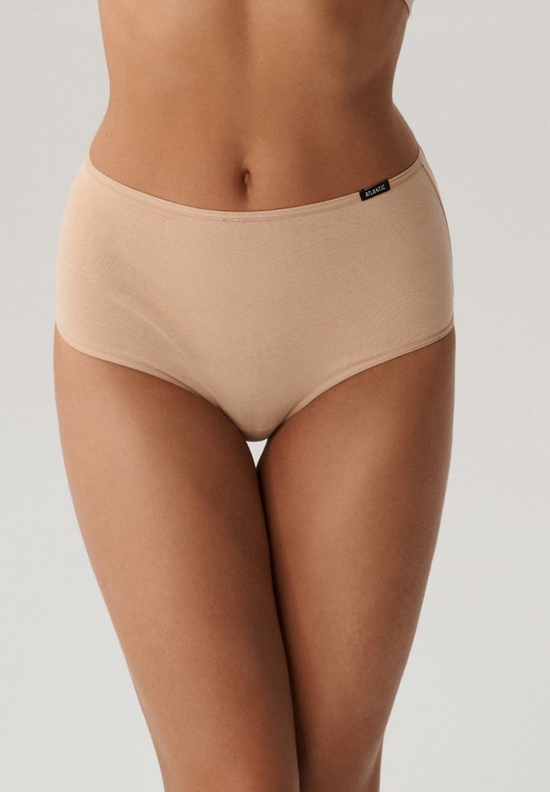 Kobieta w beżowej, wysokiej, bezszwowej bieliźnie, stojąca na tle jednolitego jasnego tła.