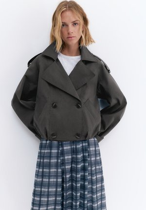 Lyshåret kvinde iført en mørkegrå dobbeltradet cropped jakke over en hvid top og et blåt ternet skørt, stående mod en hvid baggrund.