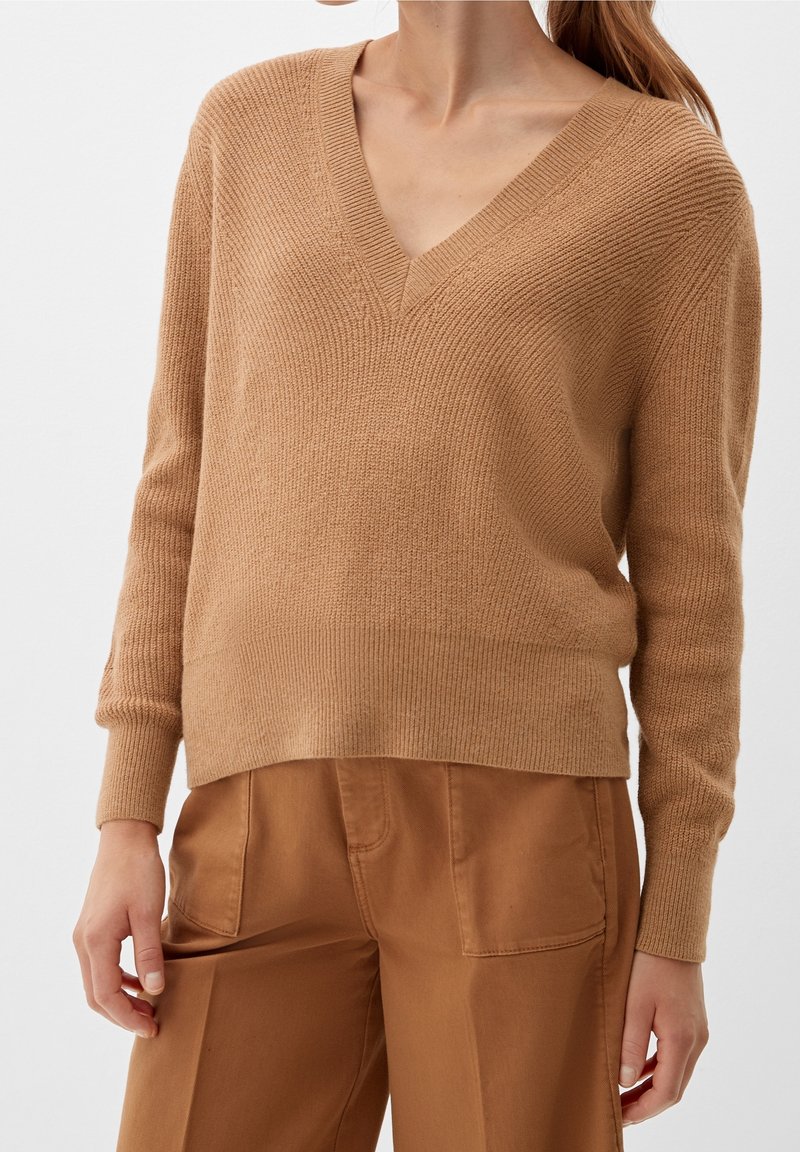 Femme portant un pull en tricot beige à col en V et un pantalon marron taille haute, debout les bras détendus le long du corps.
